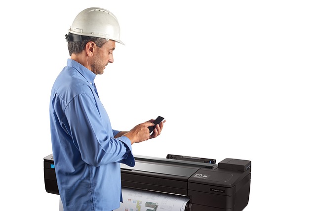 pracovník co drží mobil v ruce a velká průmyslová tiskárny co tiskne